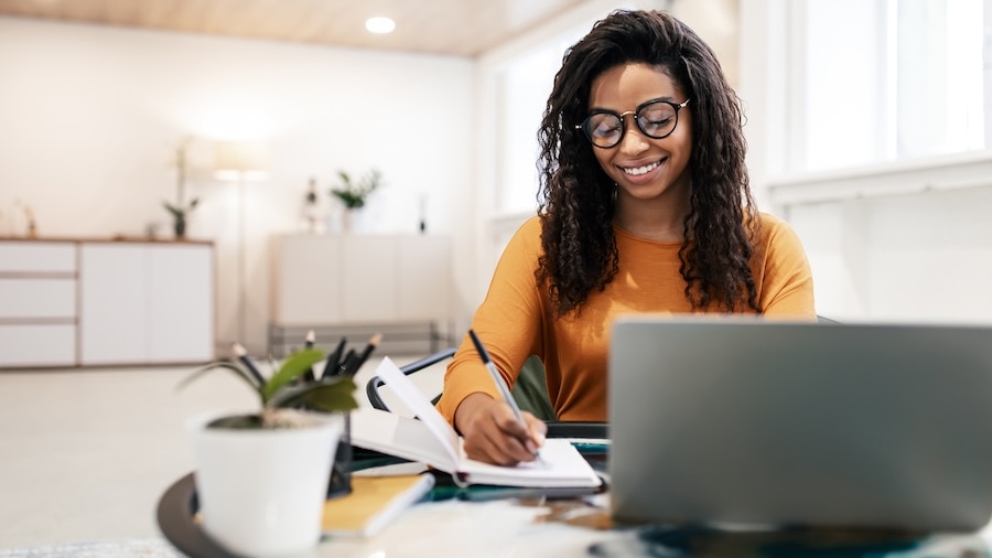 Smiling woman working from home, writing notes while using a laptop at a bright and modern workspace